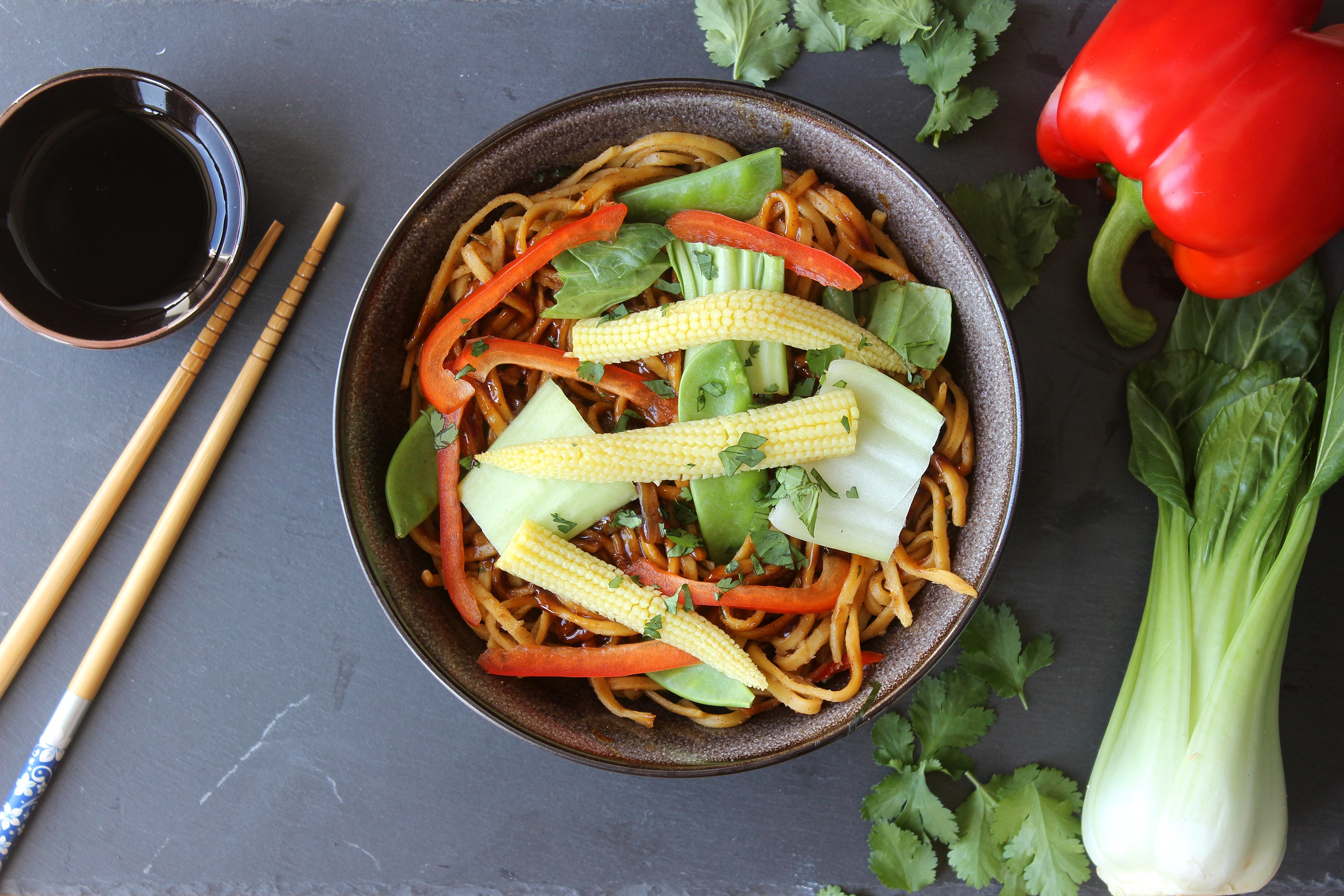 Bowl of noodles with vegetables on a dark surface with chopsticks and a red pepper.